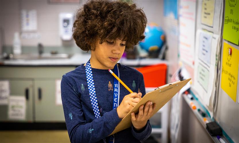 A boy wearing a blue jacket writes on a clipboard